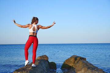 Beautiful athlete woman in red sportswear posing on the background of the sea landscape. Morning workout by the ocean, healthy lifestyle