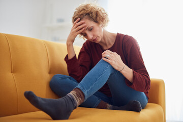 Obraz premium Woman with knee and headache problem sitting on a couch at home.