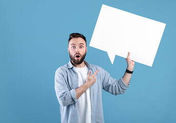 Shocked young guy pointing at empty speech bubble with space for your design over blue studio...