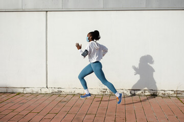 Side view of fit sporty woman running on asphalt wearing face mask against coronavirus Covid-19. Female athlete training outside under pandemic.
