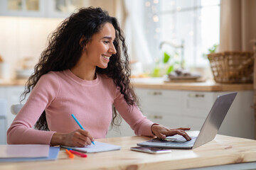 Smiling Young Freelancer Woman Working With Laptop In Kitchen And Taking Notes