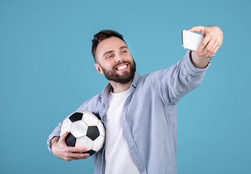Positive millennial guy with soccer ball taking selfie on smartphone over blue studio background