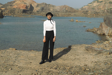 Long shot of an indian girl standing near a lake surrounded with mountains, wearing white top and black pants.