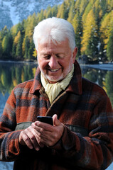 Uomo anziano digita un messaggio sul telefono cellulare  sorridente in riva al lago