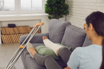 Patient with physical injury sitting on sofa at home with crutches beside. Woman with broken leg in...