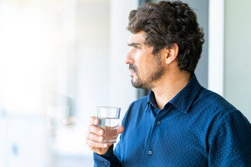 Casual businessman holding and drinking glass of water. Healthy man at home office portrait looking throught the window