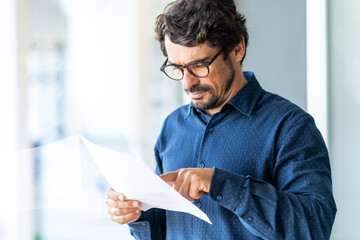 Business man wearing glasses  reading paper document or contract. Successful male portrait at the office by the window