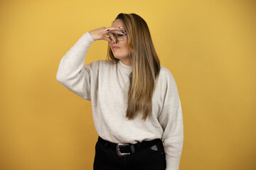 Pretty woman with long hair smelling something stinky and disgusting, intolerable smell, holding breath with fingers on nose against yellow wall