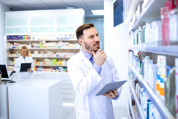 Professional handsome male pharmacist in white coat holding tablet and thinking in pharmacy store or drugstore. Healthcare and apothecary.