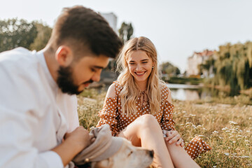Pretty blonde girl and her kind husband are resting on grass and playing with their pet. Couple posing against backdrop of lake