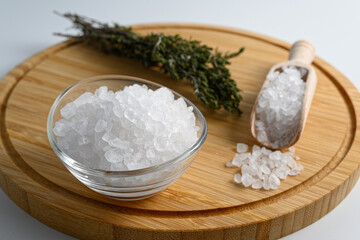 Little wooden scoop and glass bowl full of sea salt on wooden cutting board on white background