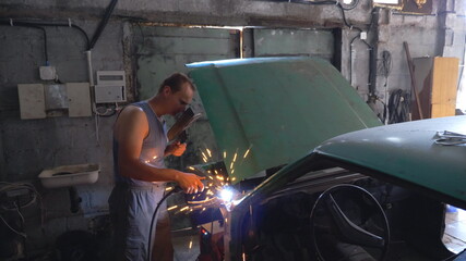 Professional repairer wearing protective mask welding auto details. Mechanic working in garage or workshop. Man engaged servicing vehicle. Hot sparks flying around. Car repair and maintenance concept