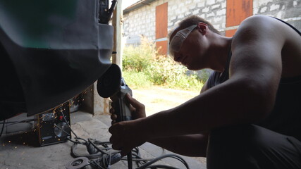 Mechanic wearing protective glasses polishing auto with electric tool in garage. Skillful mechanic restoring automobile. Guy grinding body of old car using grinder machine. Man engaged servicing car