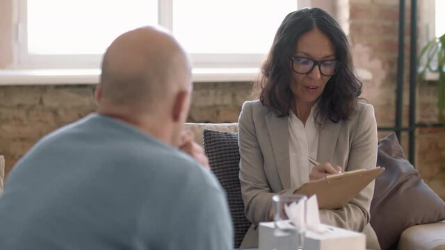 Tracking shot of female psychotherapist in glasses writing notes on clipboard and talking to emotional male client during therapy session in her private practice