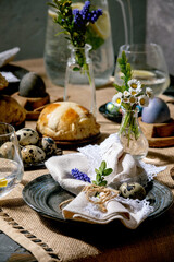 Easter table setting with colored and chocolate eggs, hot cross buns, bouquet flowers, empty ceramic plate with napkin, glass of lemonade drink on wooden table with textile tablecloth.
