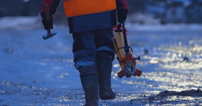 Excavation at a construction site, a surveyor with a tripod and a hammer walks on a mud road of a construction site to another point for measurements 