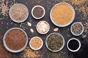 Healthy seeds - sesame, flax seed, sunflower seeds, pumpkin seed, chia and black seed in bowls on a black stone background. Top view
