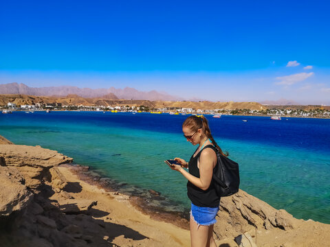 Young caucasian woman tourist looks at a photo on a smartphone screen in Sharm El Sheikh (Egypt). Female person in summer clothes on the background of the seascape of the Sharm el-Maya bay