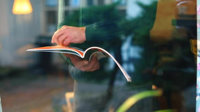 A man leafs through a magazine near a window in a self-isolation house. He turns the page with his hand. Use of printed publications on paper. Camera movement through glass, glare, reflections.
