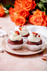 Homemade Red velvet cupcakes with whipped cream on pink ceramic plate, white napkin with ribbon, roses flowers, wooden hearts over pink texture background. Valentines day dessert.