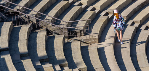 Young red-haired white woman in dress and hat seen from behind walking on the steps of a Roman...