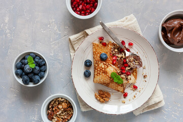 Thin pancakes on plate with pomegranate, blueberries, chocolate and walnuts on light background. Top view, close-up