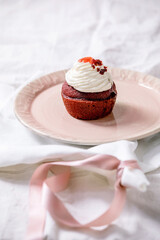 Homemade Red velvet cupcake with whipped cream on pink ceramic plate, white napkin with ribbon on white linen table cloth. Copy space