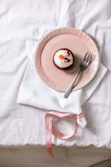 Homemade Red velvet cupcake with whipped cream on pink ceramic plate, white napkin with ribbon on white linen table cloth. Flat lay. Copy space