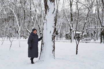 Fototapeta na wymiar Aged smiling woman standing near to a big tree in a snow-covered winter park. Snowy winter
