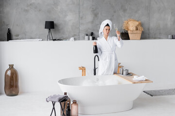 Young woman in bathrobe holding bottle and glass of champagne in bathroom
