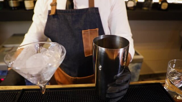 Bartender preparing a cocktail on a bar counter. Making ice with a metal tongs in a metal container with a glass of ice next to it.