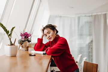 Attractive woman in stylish red outfit leaned on table. Portrait of girl sitting by window with notepad