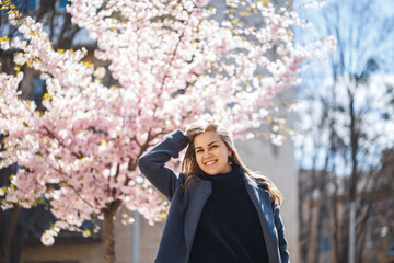 Sakura branches with flowers on a tree on the city streets. Happy woman girl in a gray palette walks along an alley with blooming sakura. Gorgeous fancy girl outdoors. Sakura tree blooming.