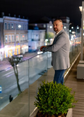 Senior elegant handsome man stands on modern balcony with glass handrail. Evening city blurred background.