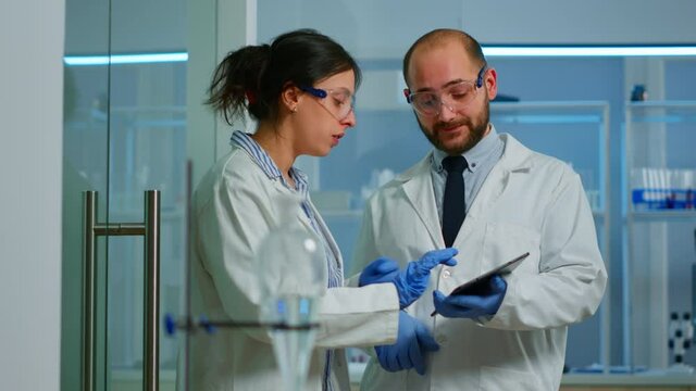 Scientist team meeting and discusssing treatment results standing in research laboratory pointing on tablet. Stuff examining vaccine development against covid19 virus using high tech for researching.
