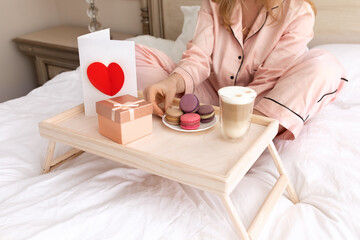 Woman on the bed with white blanket in pink cotton pajamas with a present card and a gift on Valentine's Day with coffee and sweets. Festive breakfast in bed.