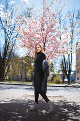 Young slender female model with long wavy hair and, dressed in a gray coat, sneakers, spinning on the street. Spring blossom trees woman girl laughs and runs to rejoice