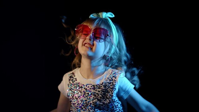 Futuristic hipster teen girl listening to music, dancing, making faces, fooling around at disco party cyberpunk night club. Cute child kid in neon blue lights camera celebrating on black background