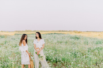 Two young unusual brunette girls in white clothes stand and pose in the middle of a blooming meadow of daisies with a bouquet of wildflowers. Enjoy the summer and nature. 