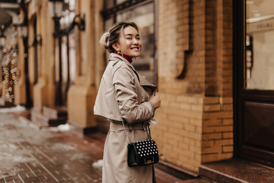 Lady with ponytail smiles and looks into camera. Portrait of blonde woman in beige trench coat and with stylish bag