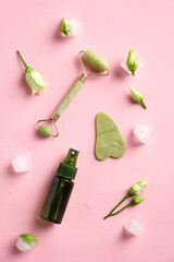 Jade stone face roller, gua sha and massage lotion on pink background with ice cubes and flowers. Skincare concept. Flat lay, top view.