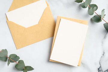 Craft paper envelopes with blank greeting cards mockups and eucalyptus leaves on marble table. Flat lay, top view.