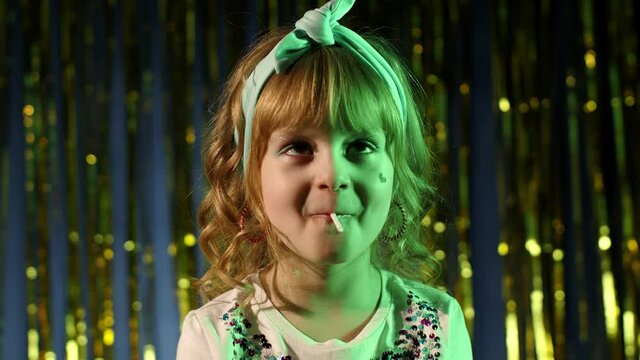Futuristic hipster teen girl licking lollipop candy in slow motion at disco party cyberpunk club. Funny caucasian child kid in neon lights standing by illuminated shiny wall. Rolled eyes of pleasure
