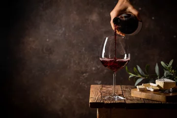 Tuinposter Pouring red wine into the glass against rustic dark wooden background © petrrgoskov
