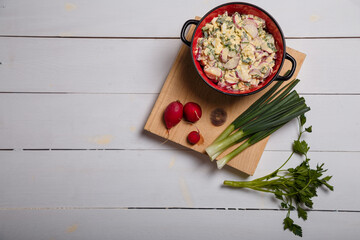 Radish, green onions, parsley, eggs salad in red bowl. Some fresh vegetables and greenery on kitchen chopping board on white wooden background. Top view, copy space.