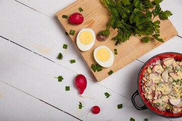 Radish, green onions, parsley, eggs salad in red bowl. Some fresh vegetables and greenery on kitchen chopping board on white wooden background. Top view, copy space.
