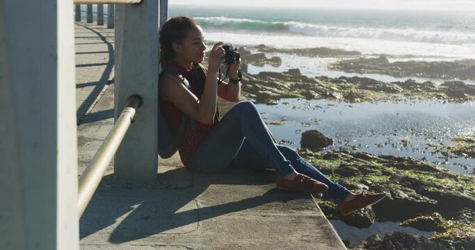 African american woman sitting and photographing on promenade by the sea