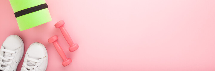 Female fitness training accessories. White sport shoes, dumbbells, green foam mat on light pink table background. Pastel color. Wide banner. Empty place for motivational, inspirational text. Top view.