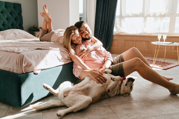 Guy and his girlfriend are resting in bedroom. Happy couple lovingly looking at their pet who wants...