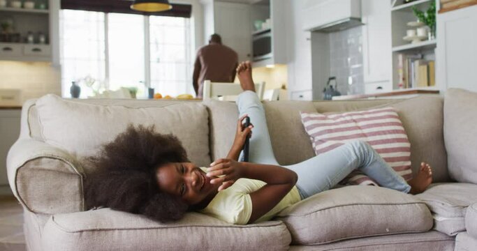 African american daughter lying on couch laughing at tv with father in background